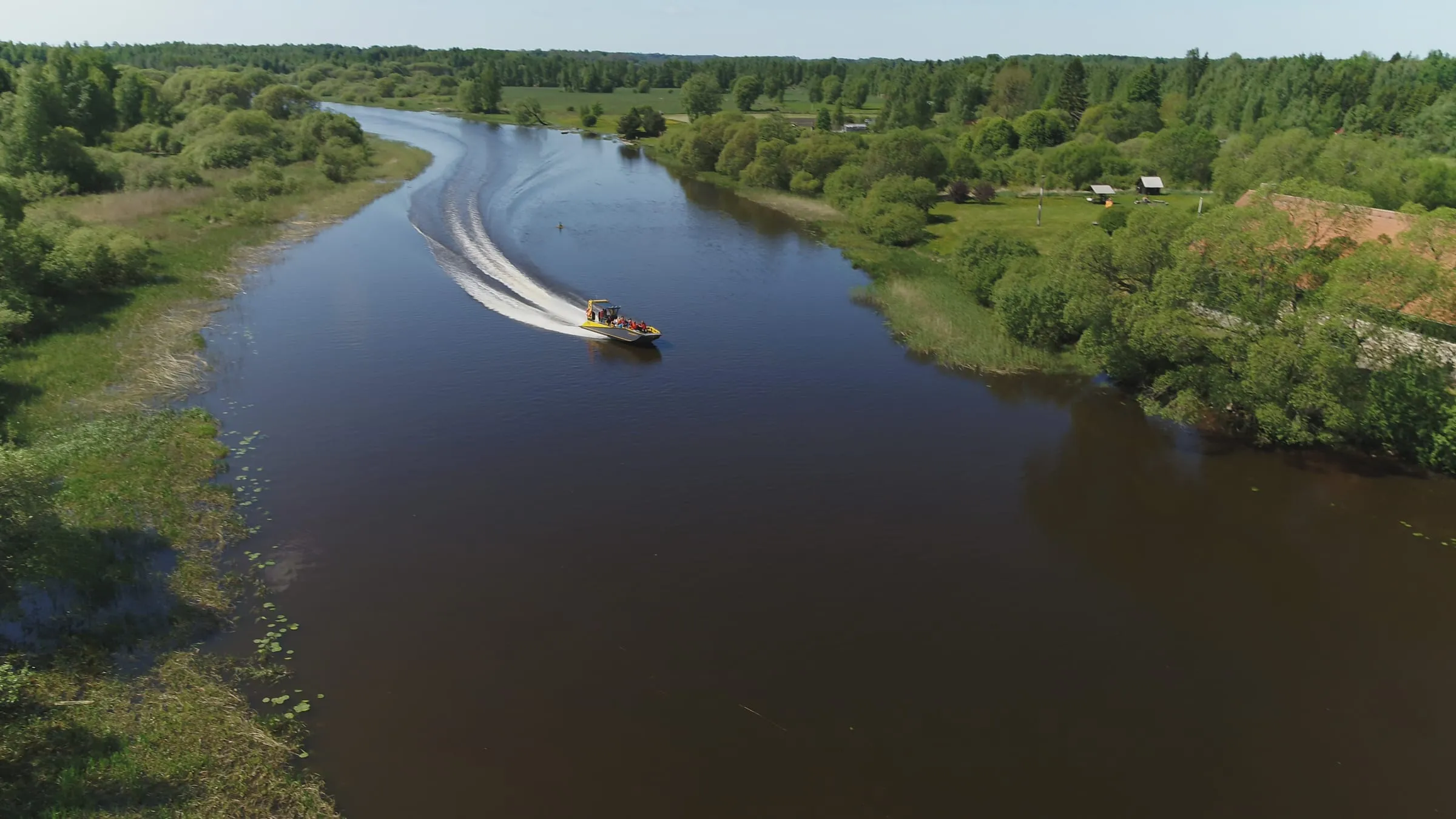 Aerial view of an LR9 Safari running a narrow, winding river between wooded banks, passengers in lifejackets on the forward benches and a long clean wake curving behind