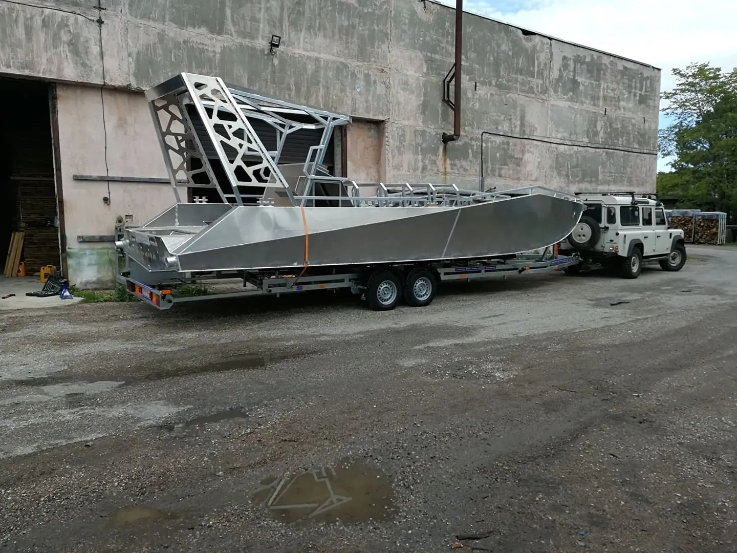 A bare, unpainted LR9 Safari hull strapped to a twin-axle road trailer outside a boat builder's hall, the aluminium wheelhouse frame welded up but not yet plated
