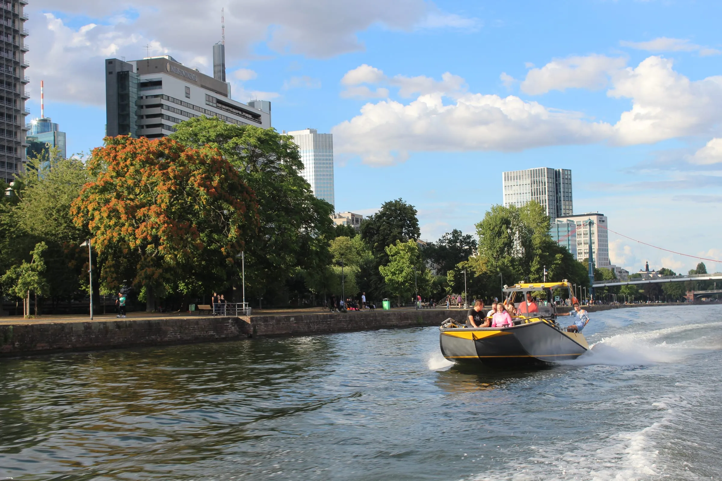 A black and yellow LR9 Safari underway on the river Main in Frankfurt, passengers on the open forward deck and one crew member at the helm behind them, the city waterfront and high-rise skyline behind