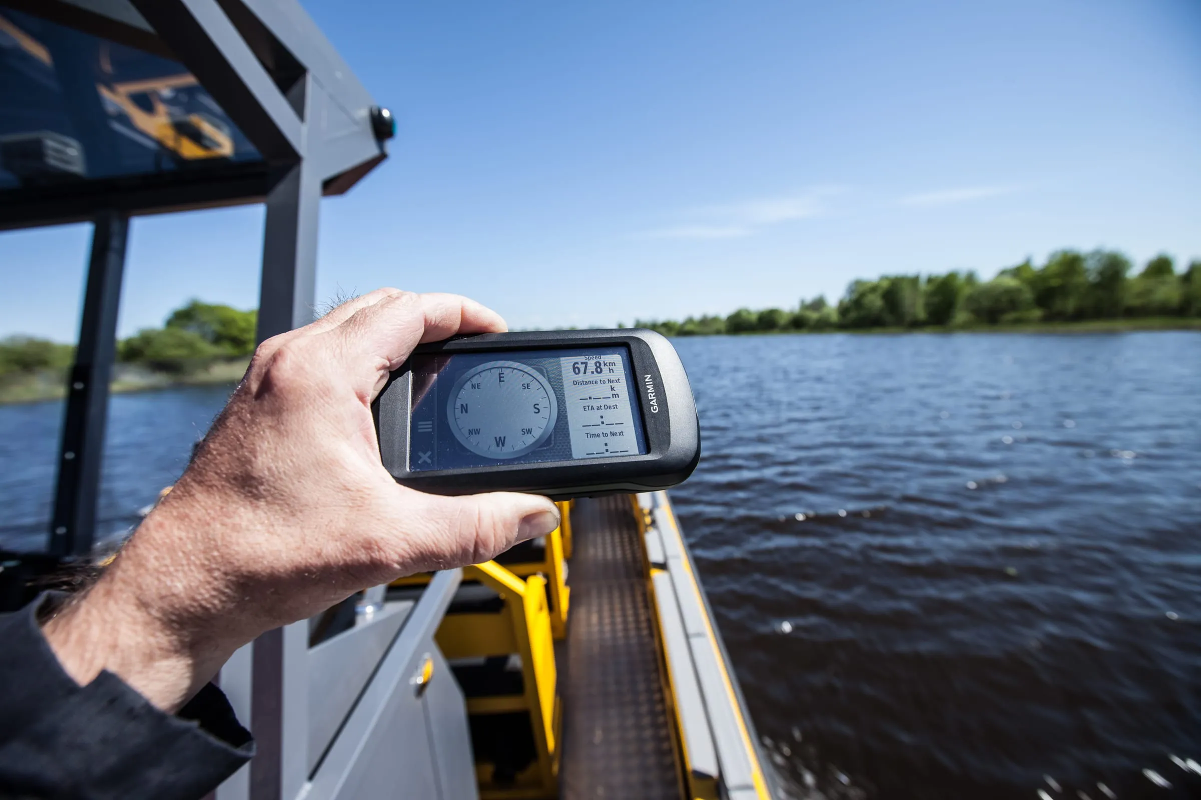 A hand holding a Garmin handheld GPS out over the yellow gunwale of an LR9 Safari underway, the screen reading a speed of 67.8 kilometres per hour, with the river bank beyond