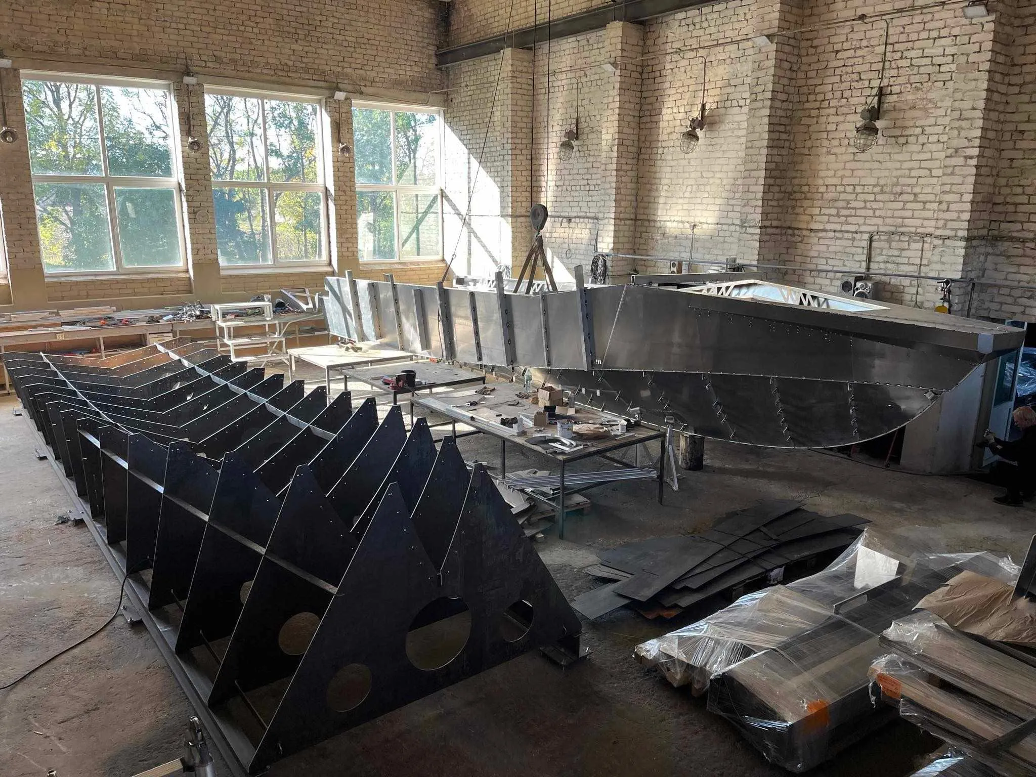An LR9 Safari hull under construction in a boat builder's hall, the plate sides already welded up and a row of cut aluminium frames standing on a jig in the foreground