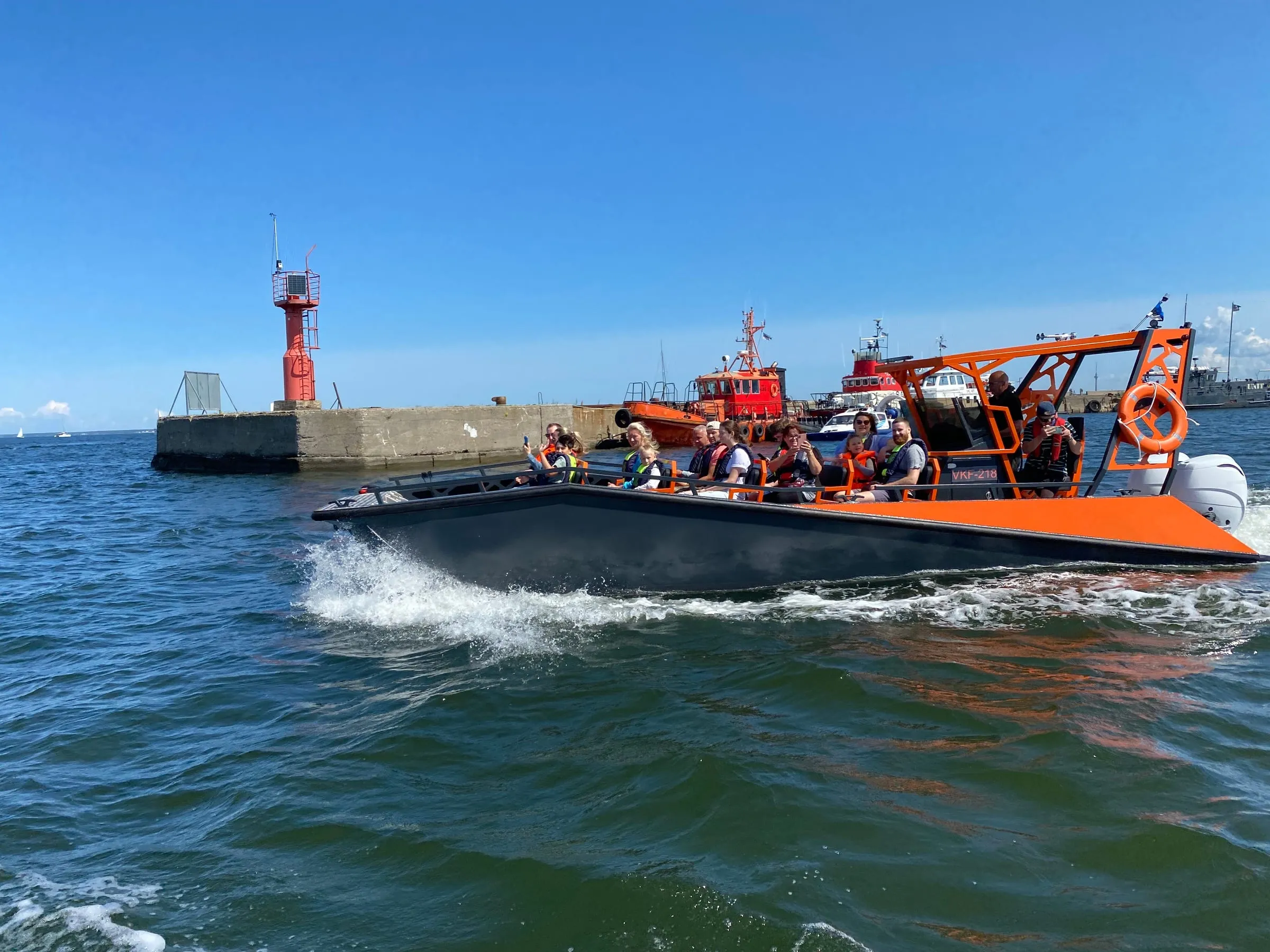An orange and grey LR9 Safari underway in a harbour entrance with about a dozen passengers in lifejackets seated on the open forward benches and two crew standing aft